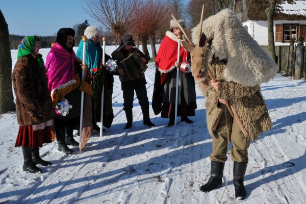 Zima, grupa przebierańców w strojach Trzech Króli, w kożuchach, na pierwszym planie chłopak w przebraniu kozy, skansen w Sierpcu
