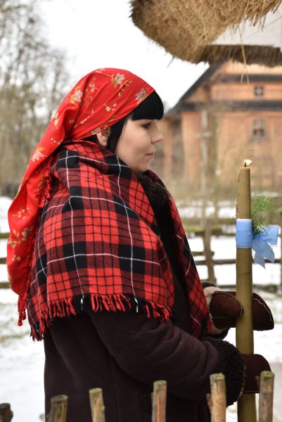 Zima, kobieta w stroju ludowym trzyma w ręku zapaloną gromnicę, wchodzi do wiejskiej chaty, w tle drewniany kościół, skansen w Sierpcu