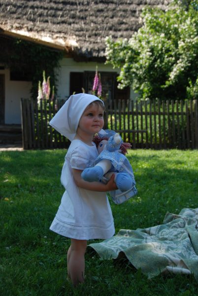 kilkuletnie dziecko w białej sukience i chusteczce na głowie trzyma zabawkę stojąc na zielonej trawie