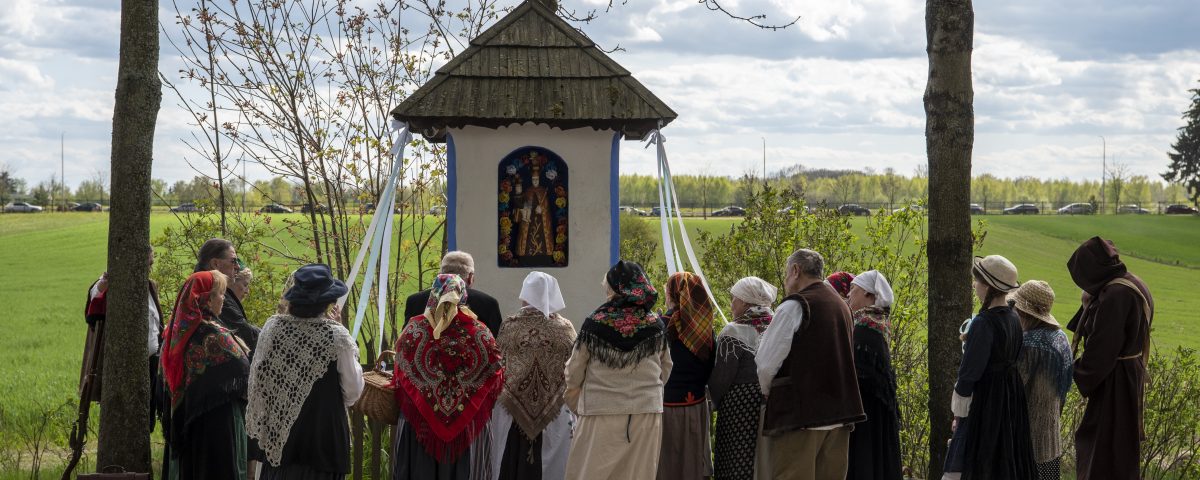 grupa osób w wiejskich strojach przy przydrożnej kapliczce modli się na nabożeństwie majowym