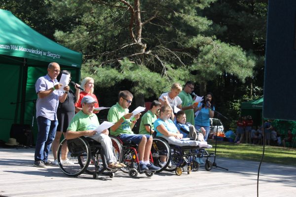 osoby niepełnosprawne na wózkach inwalidzkich oraz ich opiekunowie , w tle polana i drzewa