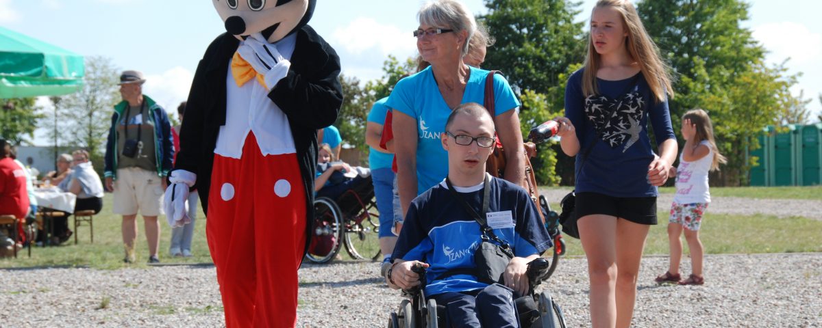 mężczyzna niepełnosprawny na wózku w centralnej części, za nim kobieta w niebieskiej koszulce, po prawej kobieta w czarnej sukience i długich blond włosach, po lewej człowiek w przebraniu Myszki Miki