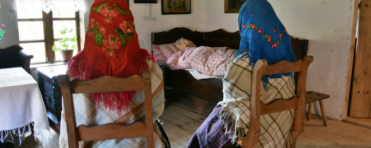 Dwie osoby siedzą na krzesłach przy łóżku, w którym leży zmarły