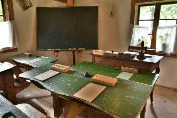 izba szkolna sprzed 100 lat. po lewej stronie tablica, zielone ławki szkolne a na nich zeszyty i kałamarze
